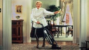 A person dressed as an older woman vacuums a rug in a living room, holding one hand out and appearing to sing or speak.