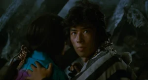A man in a striped garment looks forward while holding another person close; the background is dark and out of focus.
