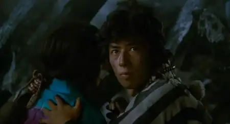A man in a striped garment looks forward while holding another person close; the background is dark and out of focus.