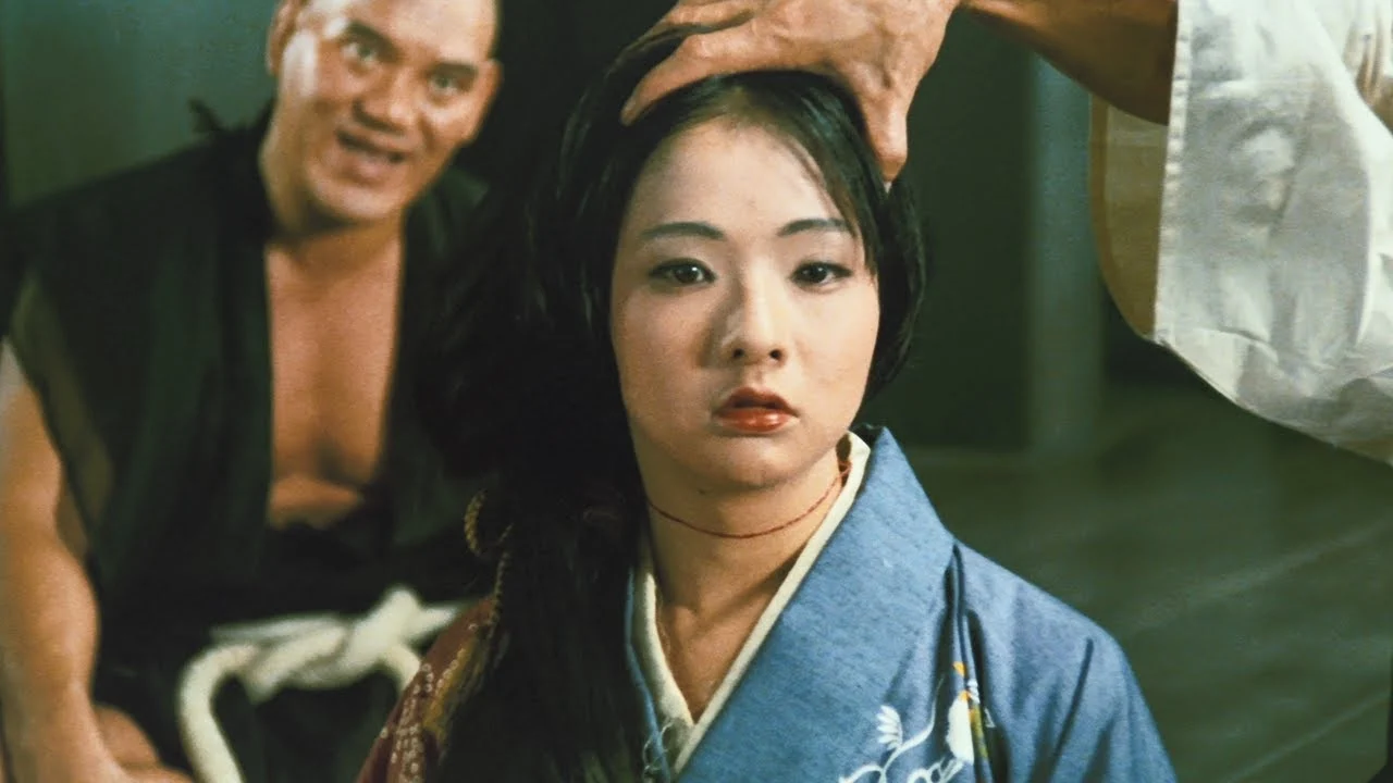 A woman in a blue kimono sits expressionless as a hand rests on her head; a man in black clothing appears in the background.