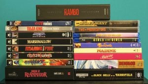 No Streaming Required: Physical Media Spotlight For The Week Of September 15th A stack of various Blu-ray and DVD cases, including titles like “RAMBO,” “Tulsa King,” “Demigod,” and “Bride of Reanimator,” arranged in two uneven piles against a teal background.