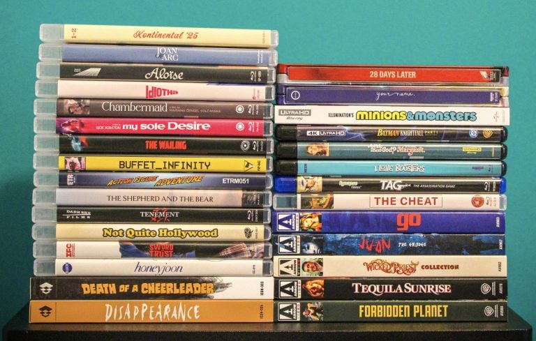 A stack of DVDs and Blu-rays stands against a teal wall, featuring various movie titles with diverse genres, arranged in two uneven vertical columns.