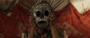 A person holds a large, ornate skull mask with metal spikes over their face; red fabric and blurred objects are in the background.