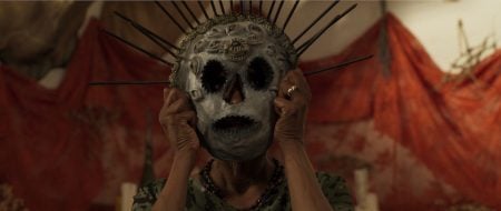 A person holds a large, ornate skull mask with metal spikes over their face; red fabric and blurred objects are in the background.