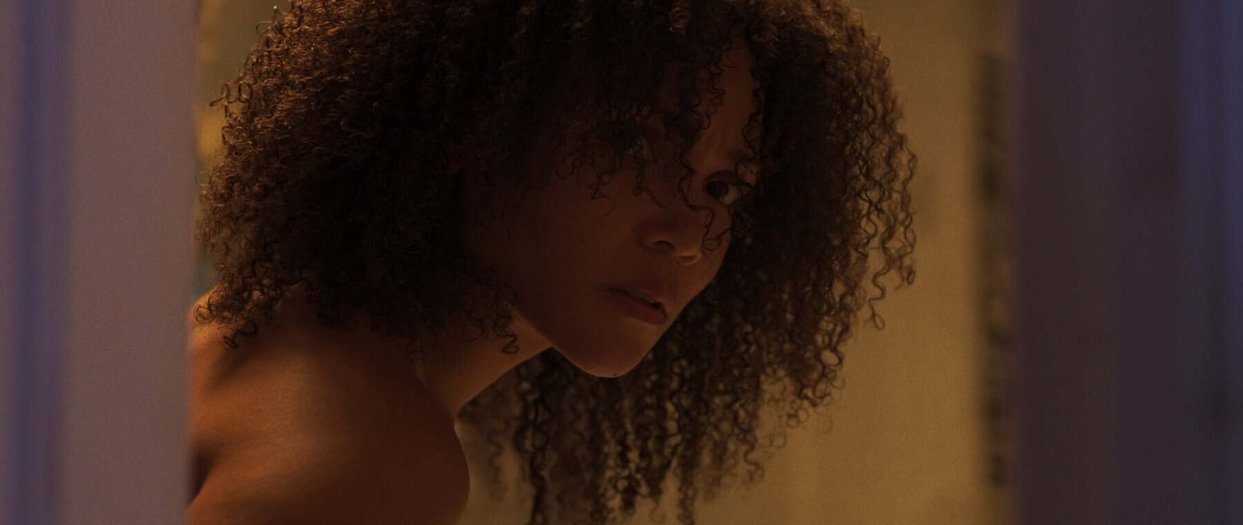 A person with curly hair looks over their shoulder while standing partially behind a door in a dimly lit room.