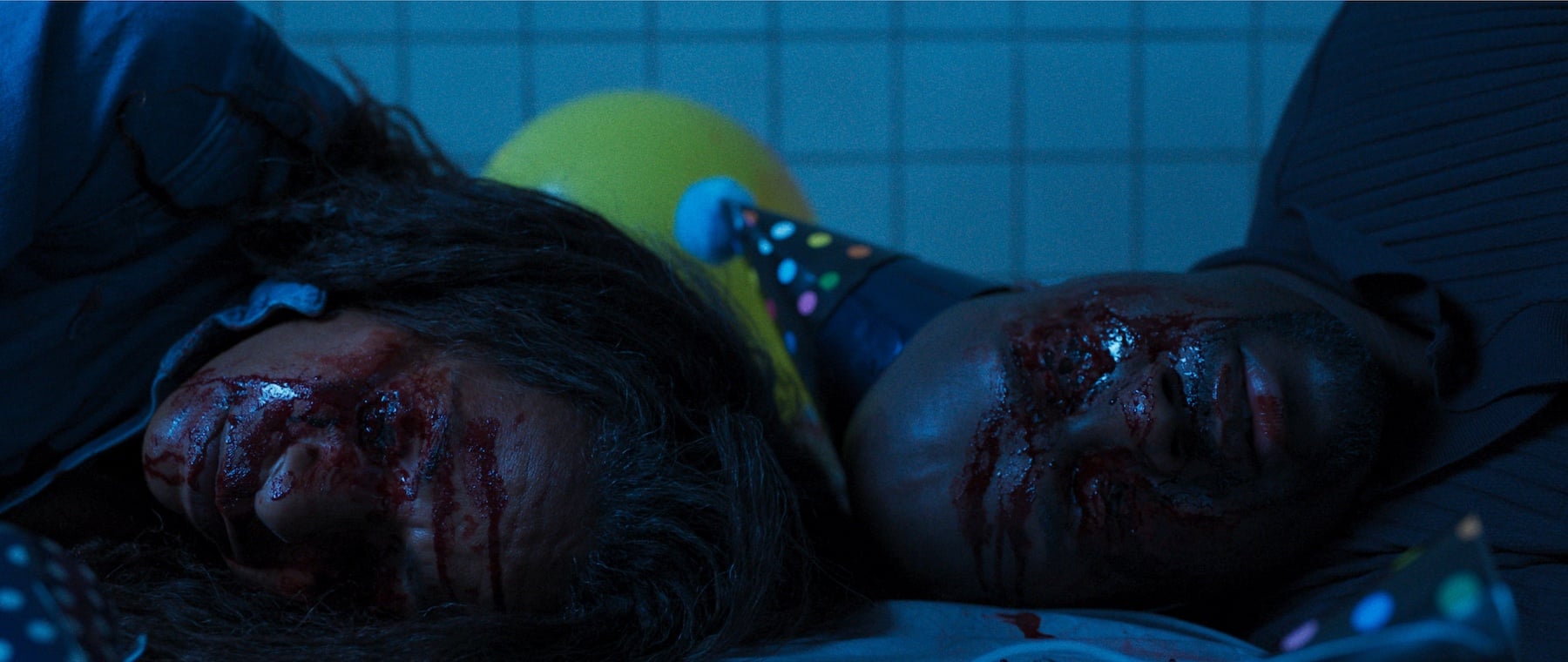 Two people with visible facial injuries and blood lie side by side on the floor, surrounded by party hats and balloons, against a tiled wall.