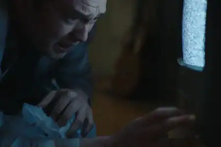 A man in a suit looks distressed as he reaches toward a small TV showing static, with crumpled tissues in his hand.