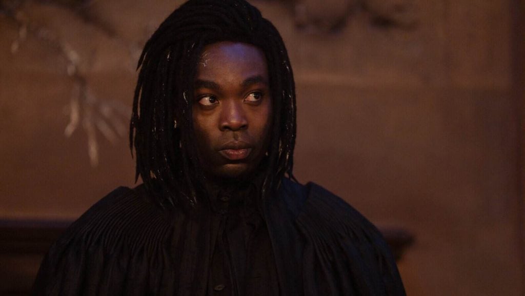 A person wearing a black robe with long, dark, braided hair stares intently to the side against a brown, dimly lit background.