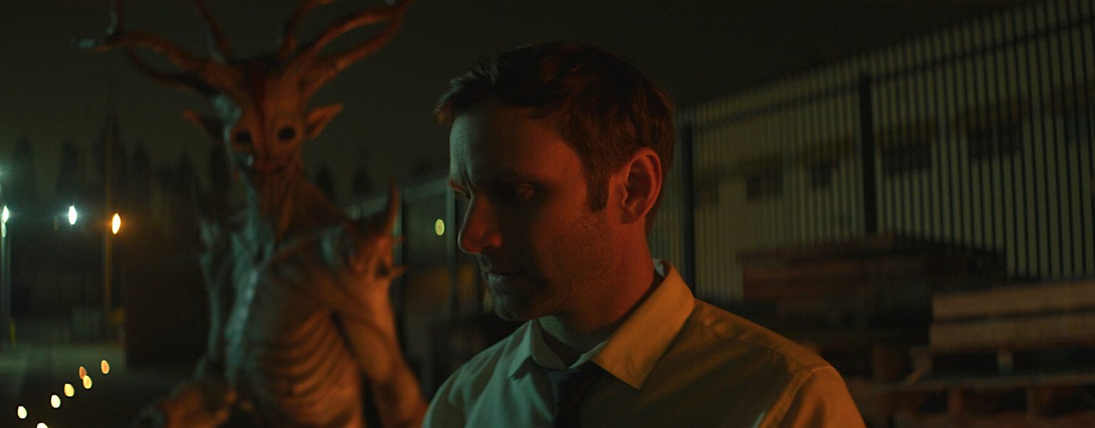 GVN Giveaway: ‘Portal To Hell’ Digital Code A man in a yellow shirt stands outside at night, looking down, while a tall, antlered creature looms behind him near a fence and wooden pallets.