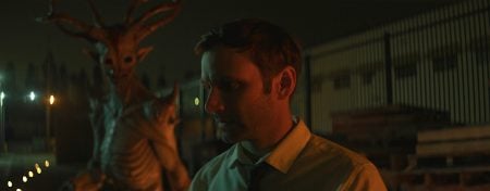 GVN Giveaway: ‘Portal To Hell’ Digital Code A man in a yellow shirt stands outside at night, looking down, while a tall, antlered creature looms behind him near a fence and wooden pallets.