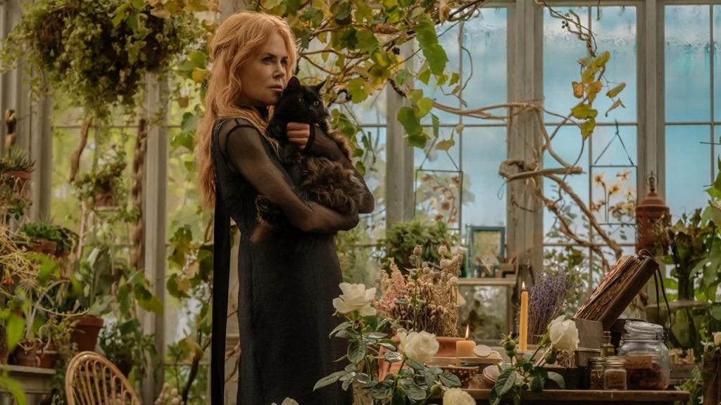 A woman with long blonde hair in a black dress stands in a greenhouse holding a black cat, surrounded by plants, flowers, and gardening supplies.