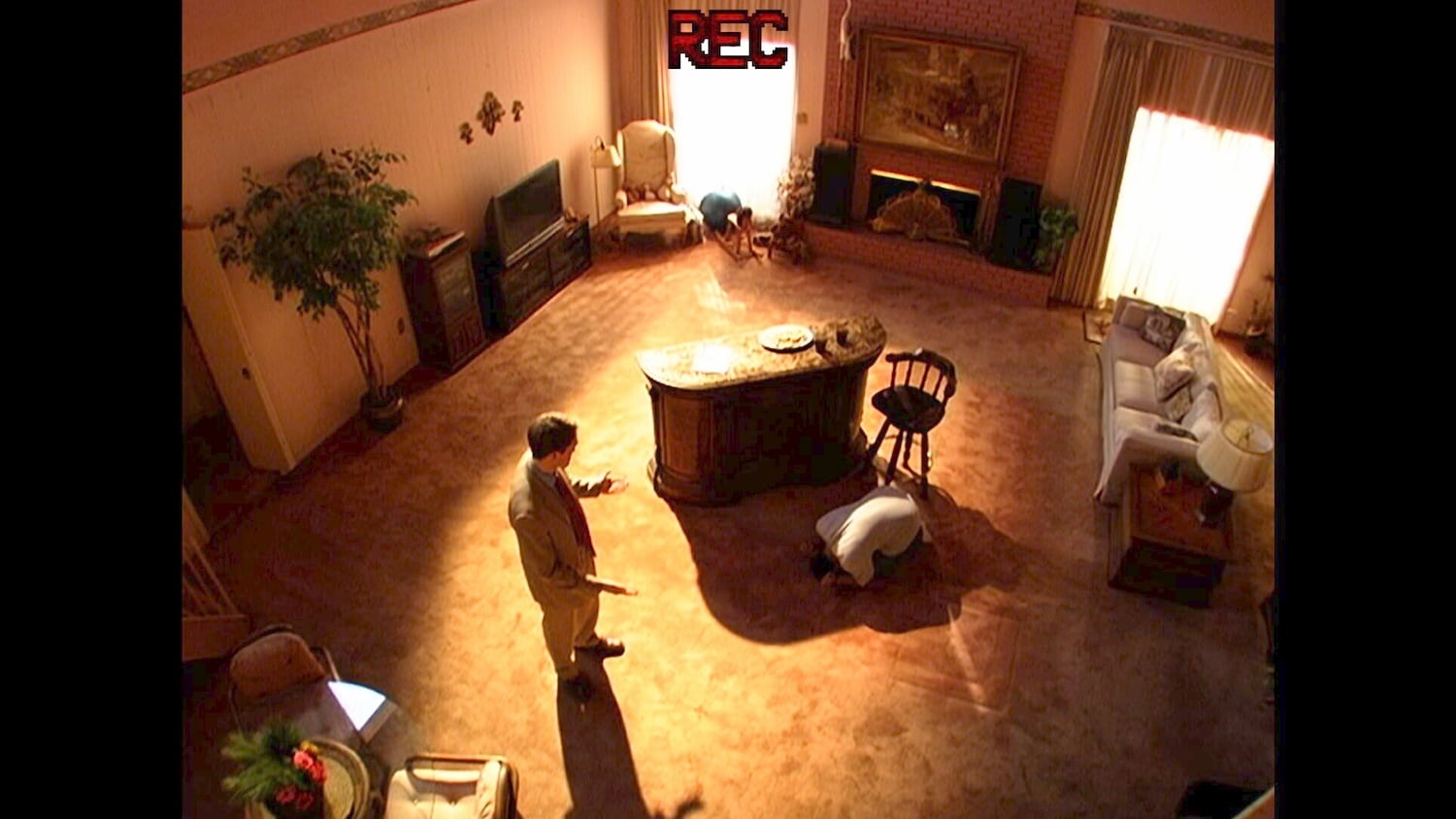A man stands in a living room near an island counter while another person kneels on the carpet. The scene is brightly lit and viewed from above, with a “REC” sign at the top of the image.