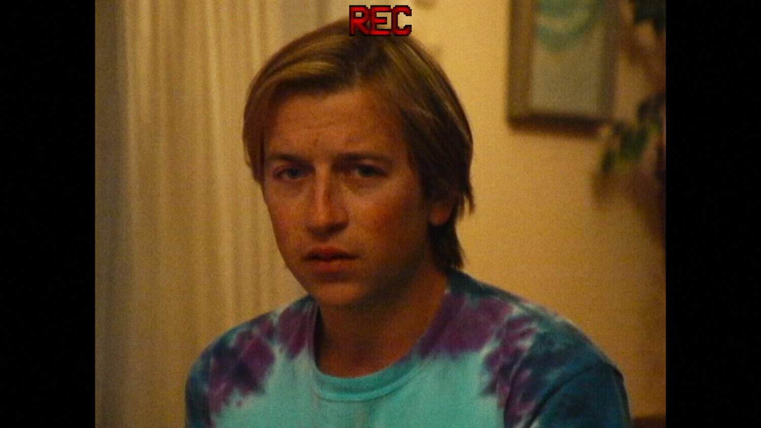 A young person with blond hair wears a tie-dye shirt and looks ahead with a neutral expression. A red “REC” icon appears at the top, indicating video recording. Indoor setting with soft lighting.
