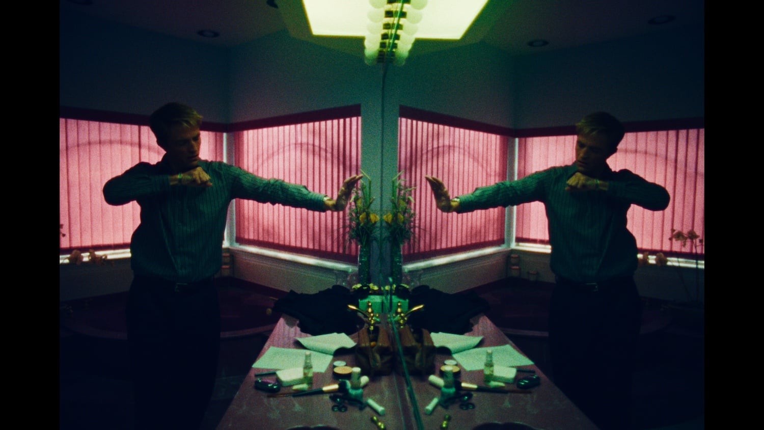 A person stands in front of a bathroom mirror with pink blinds, creating a mirrored reflection effect. Various toiletries and papers are scattered on the sink counter.