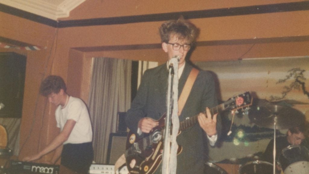 A band performs on stage; the singer plays guitar and sings into a microphone while another member plays a keyboard and a drummer is behind them.