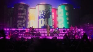 ‘Pulp: What Do You Do For An Encore?’ Review: Part Concert Film, Part Documentary, 100% Pure Nostalgia And Fun A band performs on stage with large colorful cans and the word “pulp” displayed on a screen behind them, illuminated by purple lighting.