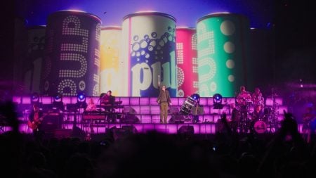 A band performs on stage with large colorful cans and the word “pulp” displayed on a screen behind them, illuminated by purple lighting.