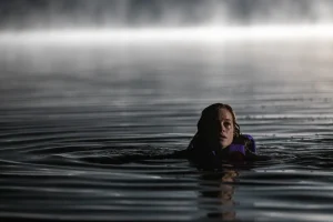 A person wearing a life jacket floats in dark, rippling water at night, with mist and light visible in the background.
