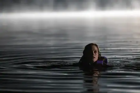 A person wearing a life jacket floats in dark, rippling water at night, with mist and light visible in the background.
