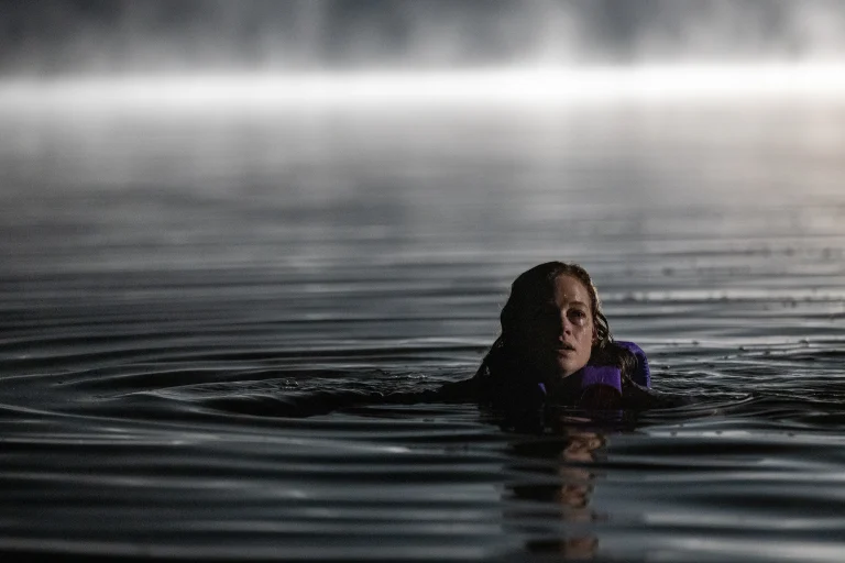 A person wearing a life jacket floats in dark, rippling water at night, with mist and light visible in the background.