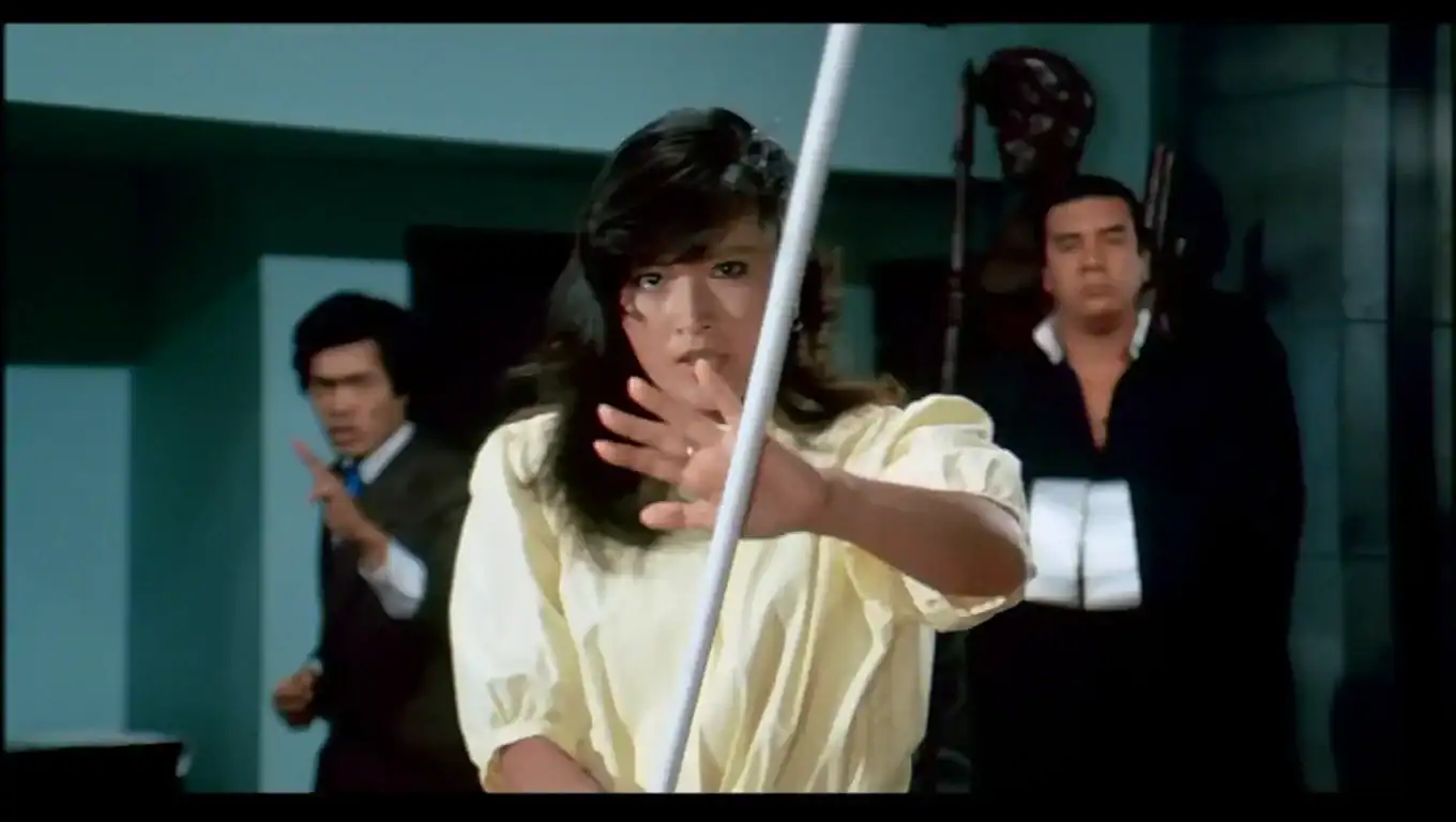 A woman in a yellow shirt blocks a sword with her hand while two men stand in the background, one pointing and one watching.