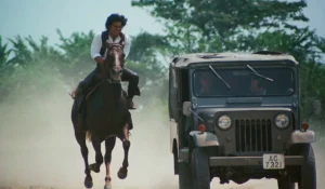 A man riding a horse at speed is parallel to a moving jeep on a dusty road, with trees visible in the background.