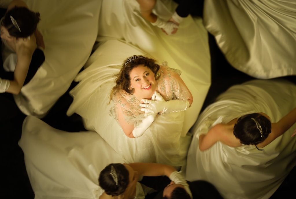 A woman in a white gown and gloves smiles up at the camera, surrounded by others in similar dresses, viewed from above.