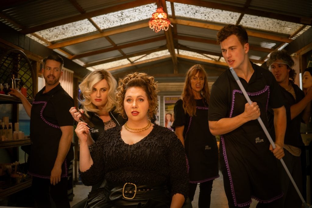 Six people wearing black salon uniforms pose dramatically inside a rustic hair salon, with two in front holding tools and four standing behind, all looking toward the camera.