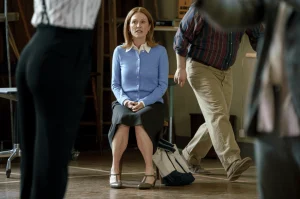 A woman in a blue sweater and black skirt sits on a chair in a room, with two people standing or walking nearby.