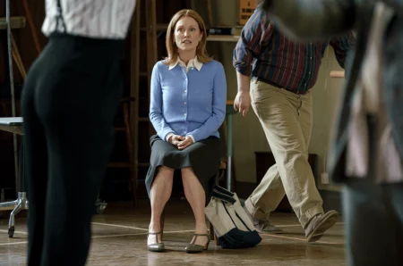 A woman in a blue sweater and black skirt sits on a chair in a room, with two people standing or walking nearby.