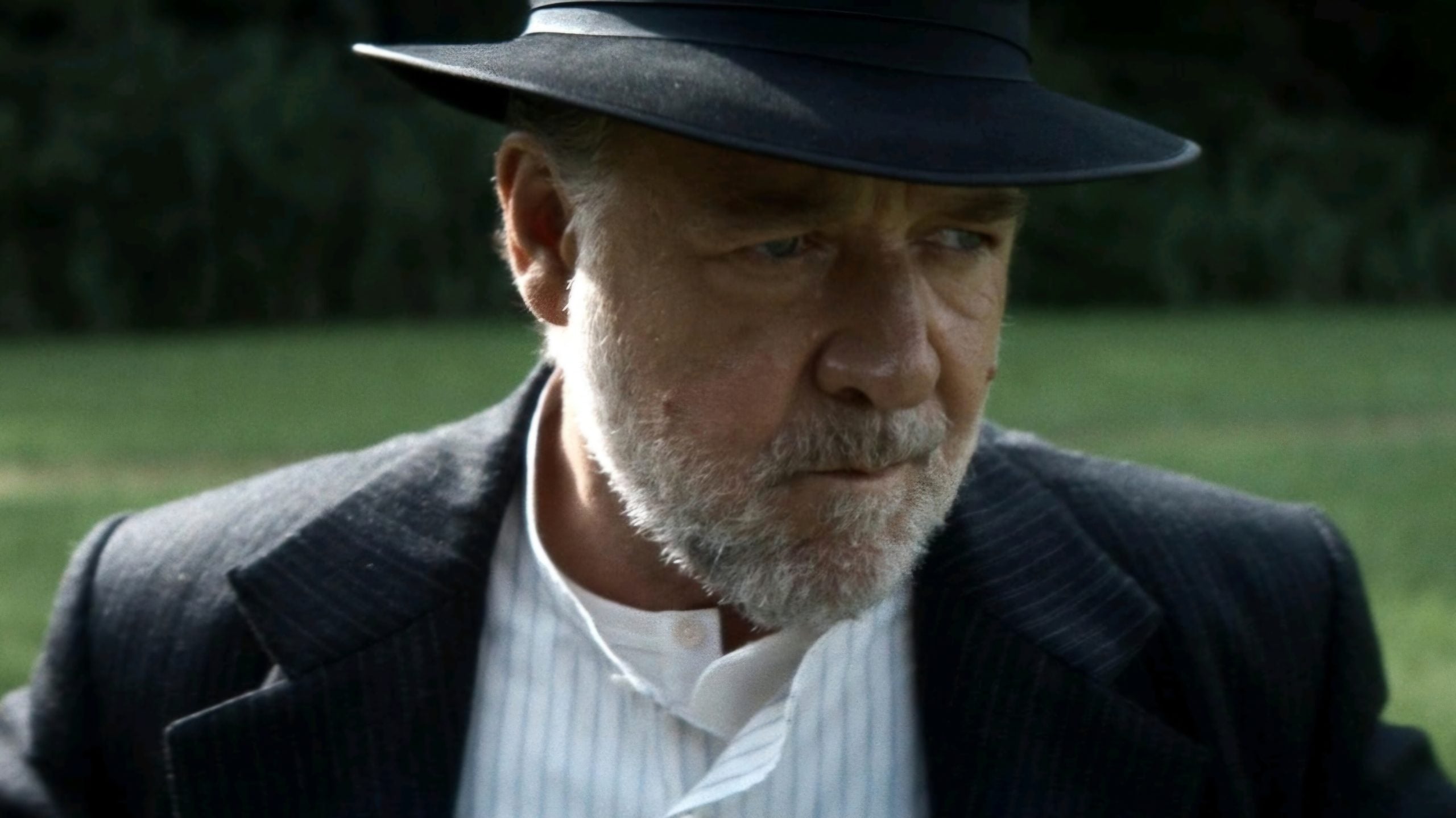 Older man with a gray beard and mustache wearing a black hat and pinstripe suit gazes downward outdoors, with greenery in the blurred background.