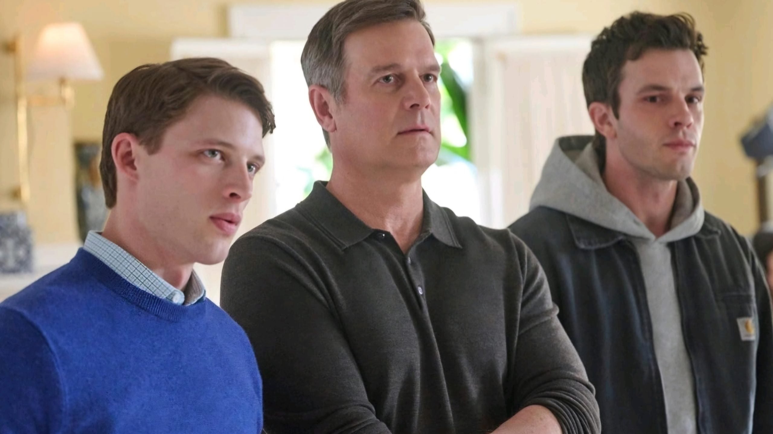 Three men standing indoors, facing forward; the man in the center wears a dark polo, while the others wear a blue sweater and a black jacket over a gray hoodie.