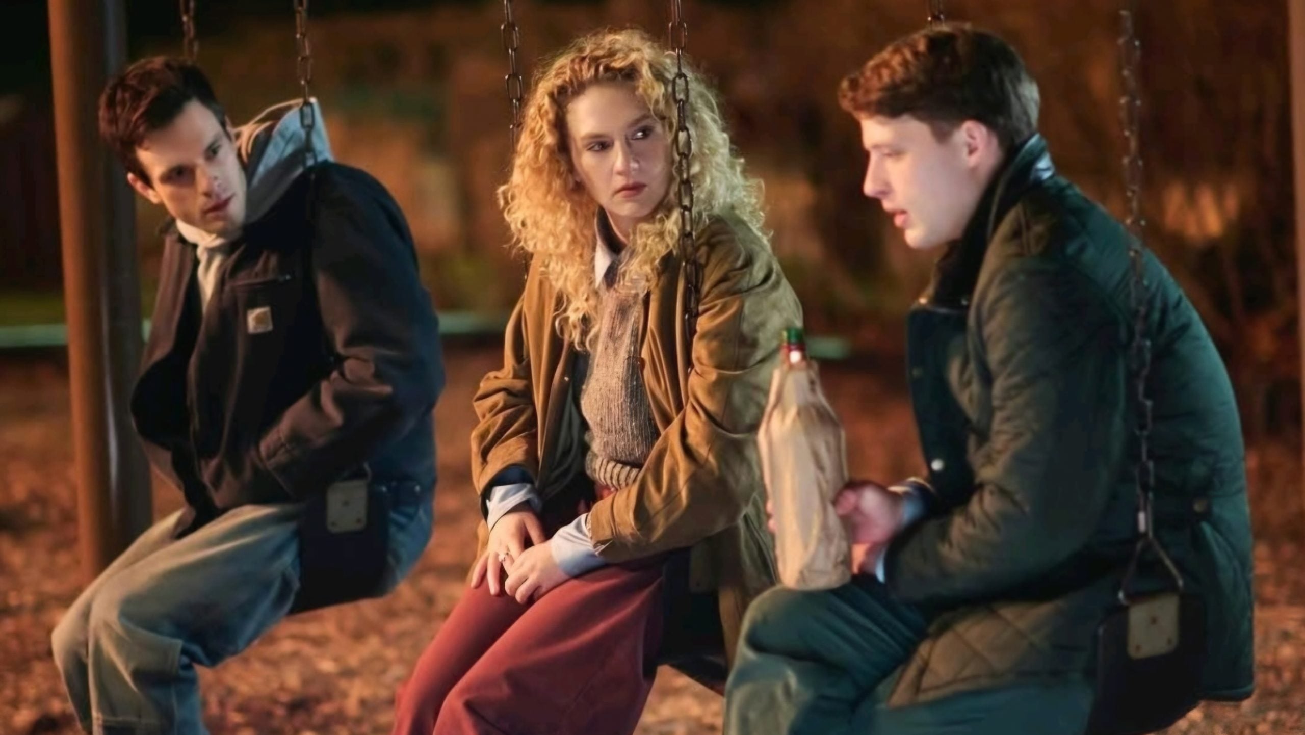 Three people sit on swings at night; the man on the right holds a brown bag bottle, and all appear to be having a serious conversation.