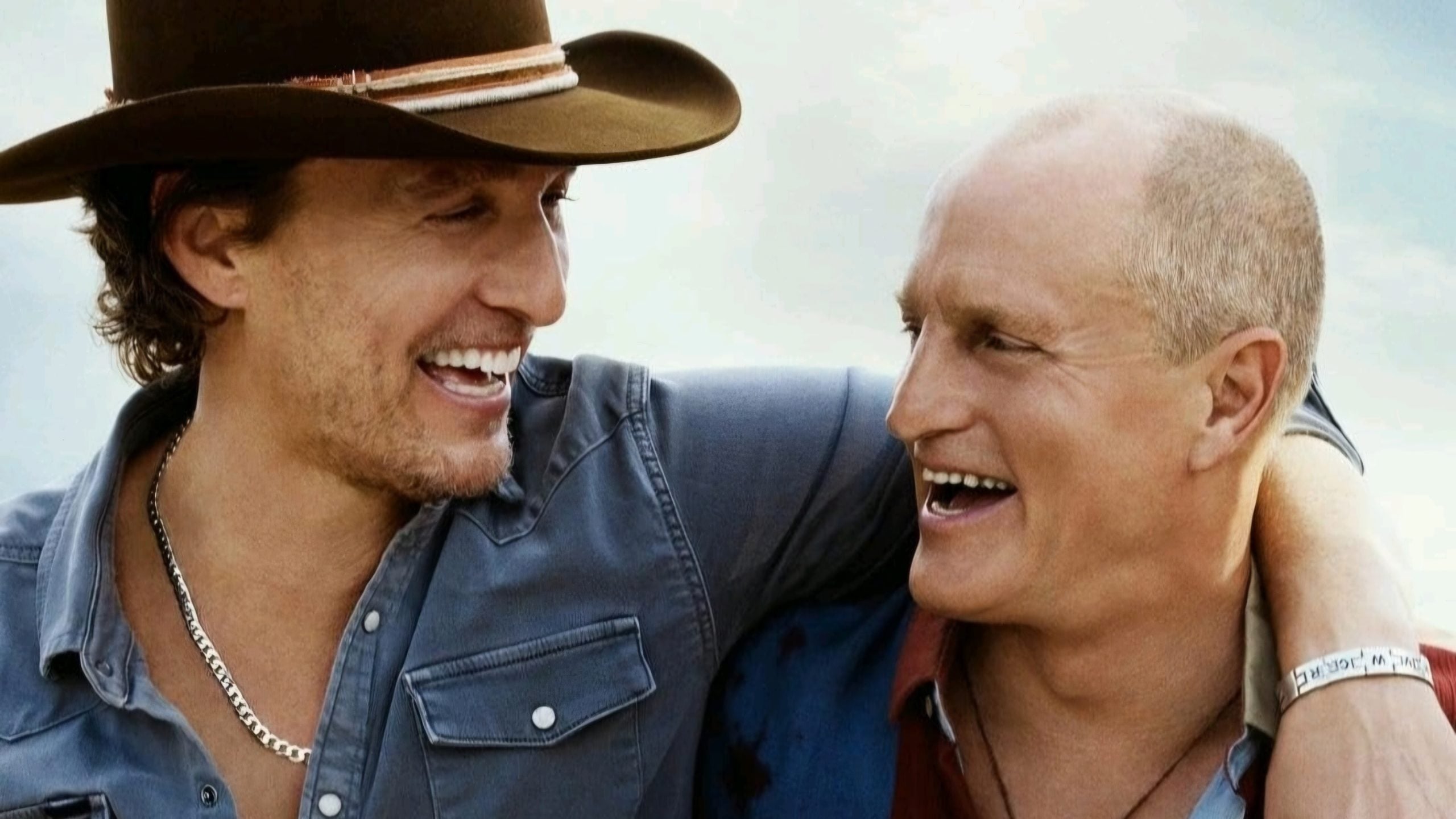 Two men smiling and laughing together, one wearing a cowboy hat and denim shirt, the other bald with a red and blue shirt, both facing each other with arms around shoulders.