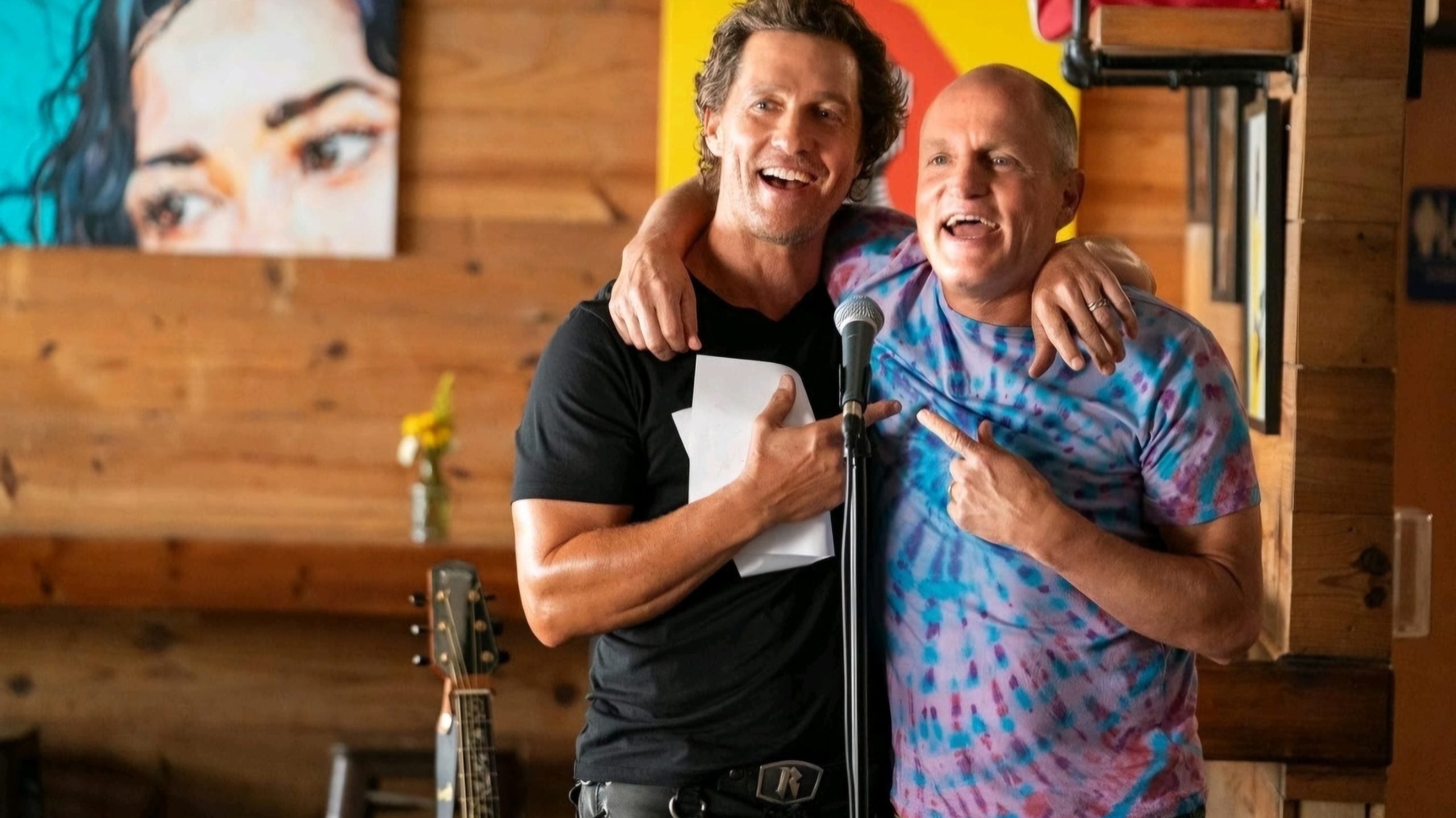 Two men stand smiling with arms around each other, both pointing at each other, in front of a microphone in a casual indoor setting. A guitar and artwork are visible in the background.