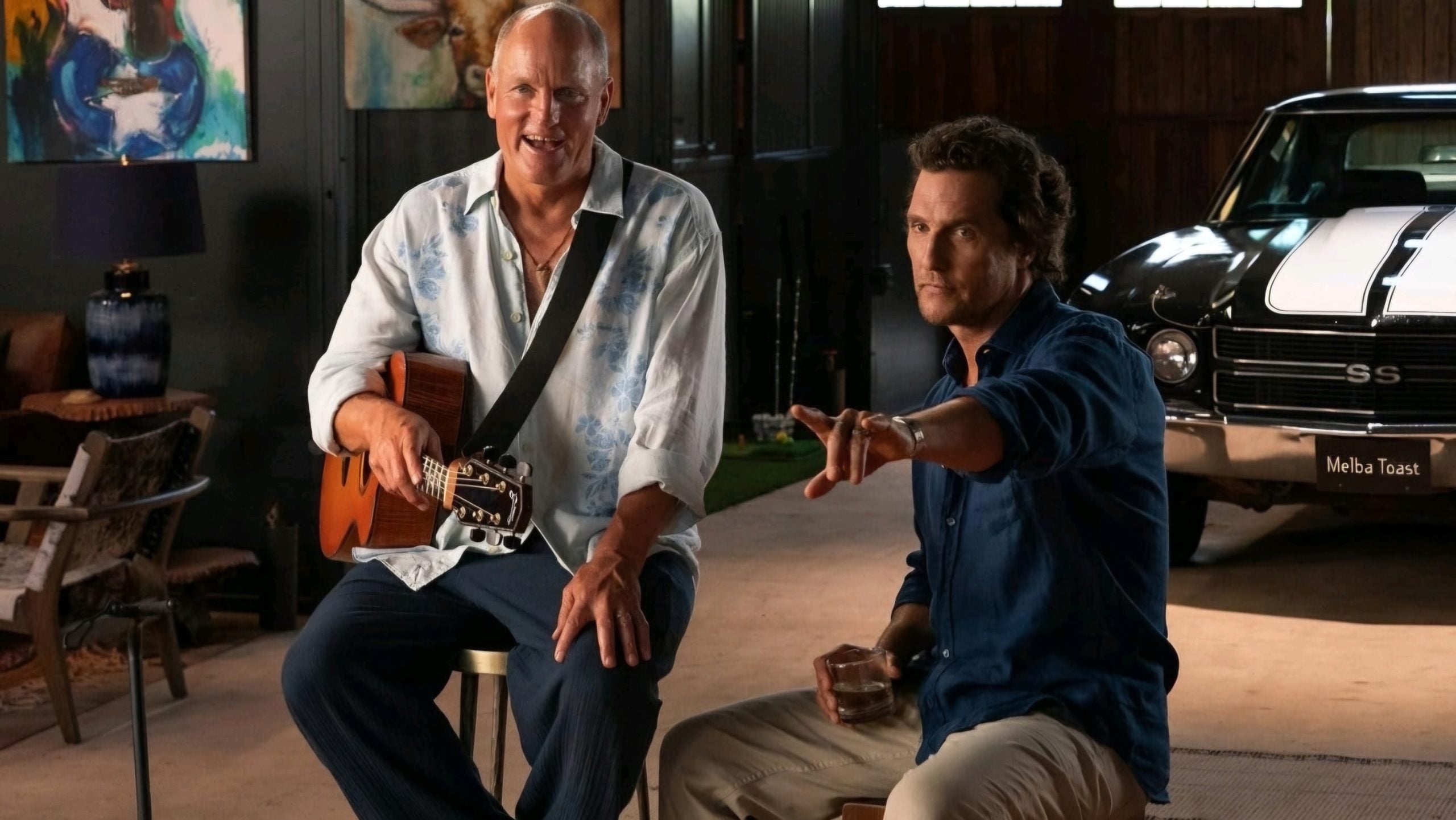 Two men sit together indoors; one holds a guitar and smiles, while the other points forward with a drink in hand. A classic car is partially visible in the background.