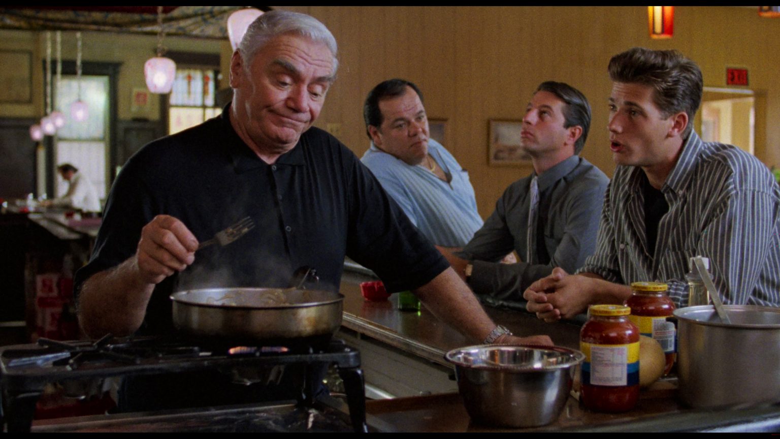 ‘Spike Of Bensonhurst’ 4K UHD Blu-Ray Review – A Genre-Blending Charmer From Paul Morrissey An older man cooks at a diner counter while three men sit and talk with him; jars of sauce and kitchen utensils are visible.