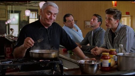 An older man cooks at a diner counter while three men sit and talk with him; jars of sauce and kitchen utensils are visible.