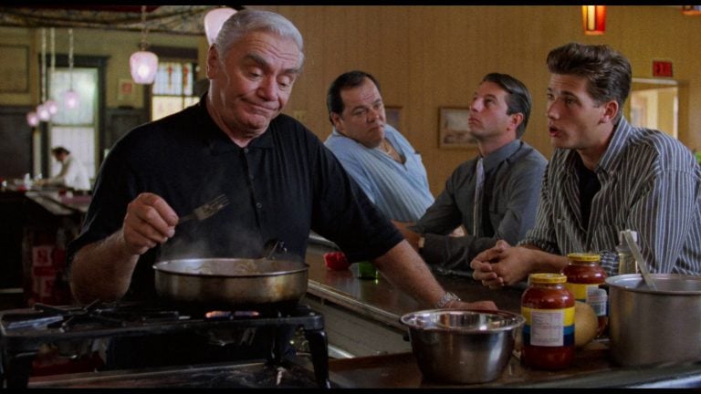 An older man cooks at a diner counter while three men sit and talk with him; jars of sauce and kitchen utensils are visible.