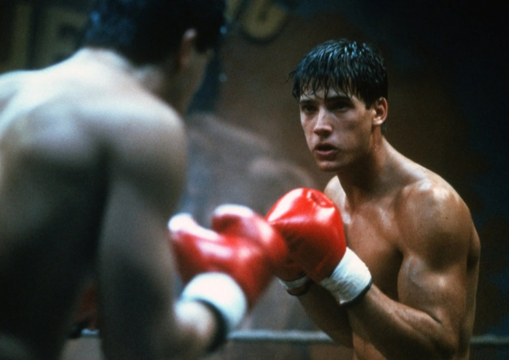 Two boxers with red gloves face each other in a boxing ring, both shirtless and in a fighting stance, with focused expressions.