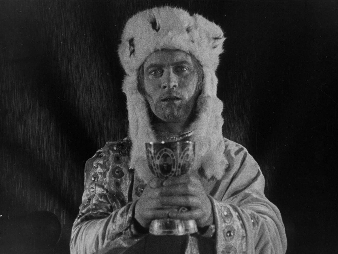 A person in ornate, medieval-style clothing and a fur hat holds a decorated chalice, looking toward the camera against a dark background.