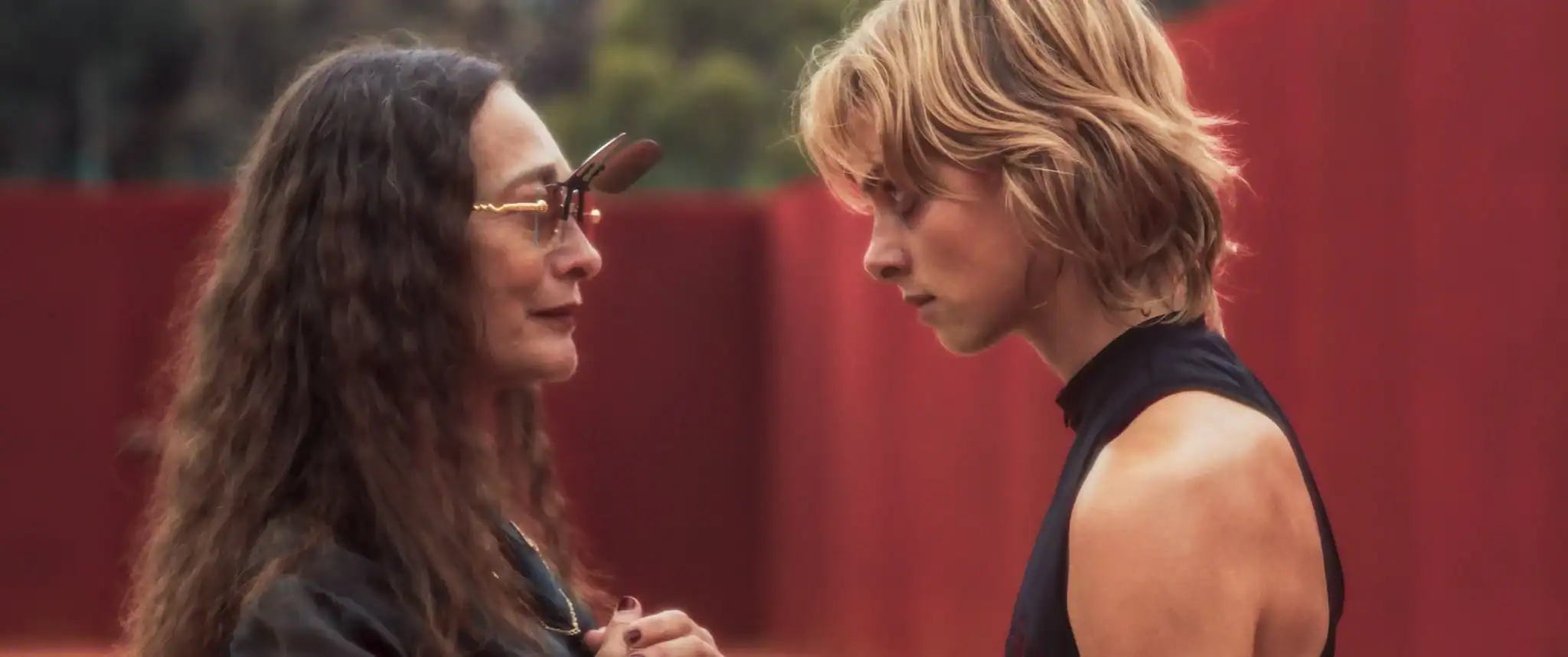 Two people stand close together outdoors, facing each other intently. One wears glasses and has long hair; the other has short blonde hair and wears a sleeveless top. A red wall is behind them.