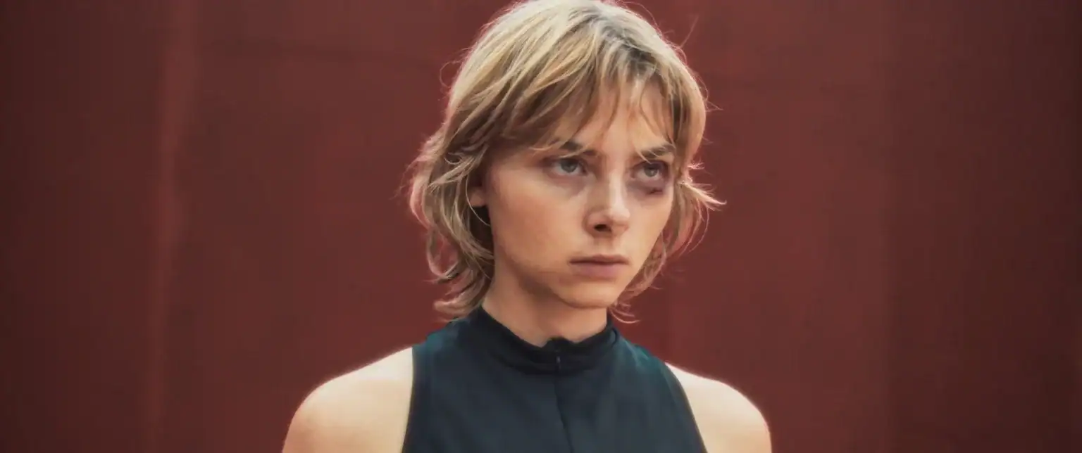 Person with short, tousled blond hair wearing a sleeveless black top stands against a plain red background, looking forward with a serious expression.