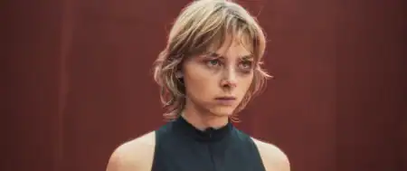 Person with short, tousled blond hair wearing a sleeveless black top stands against a plain red background, looking forward with a serious expression.