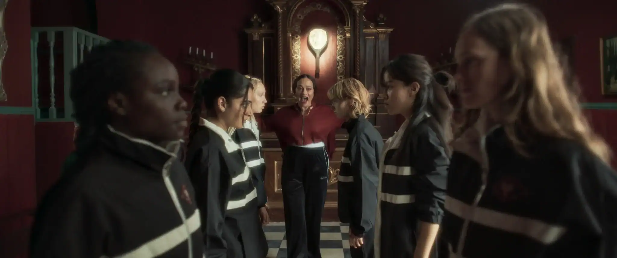Six young women in black uniforms stand in two rows facing each other, while an adult woman in red stands between them in an ornate, dimly lit room with a checkered floor.