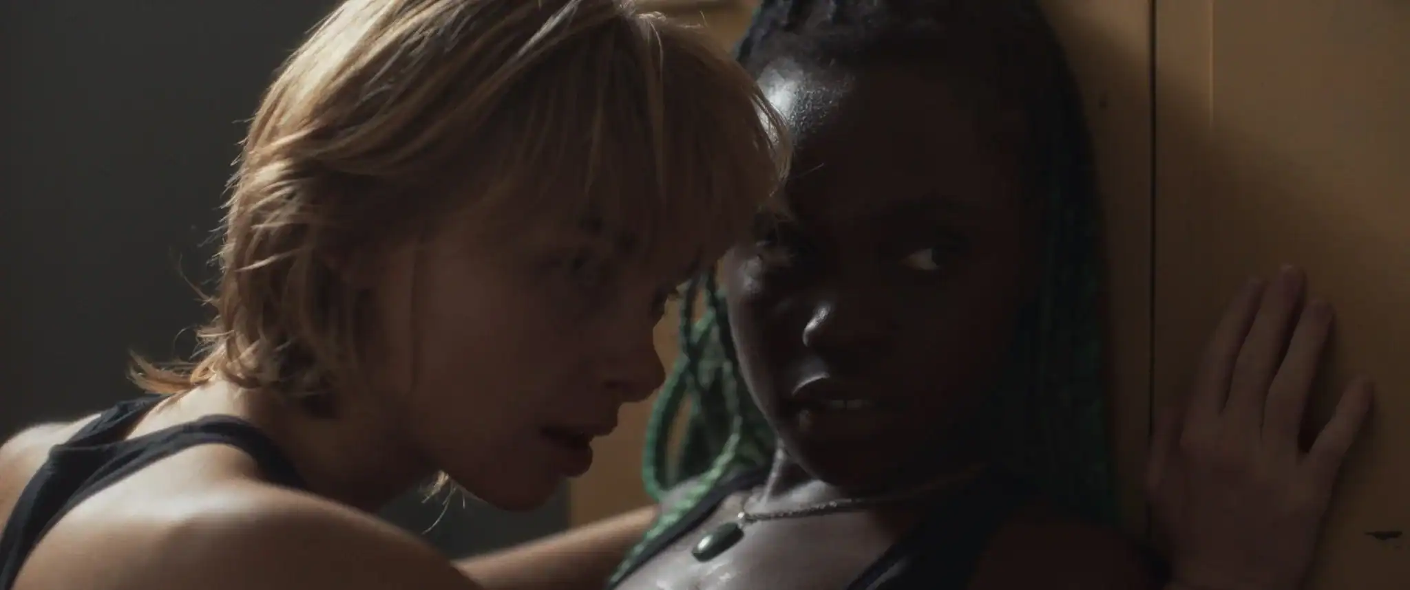 Two women standing close together indoors, one with short blonde hair and the other with long green braids, both looking in the same direction with serious expressions.
