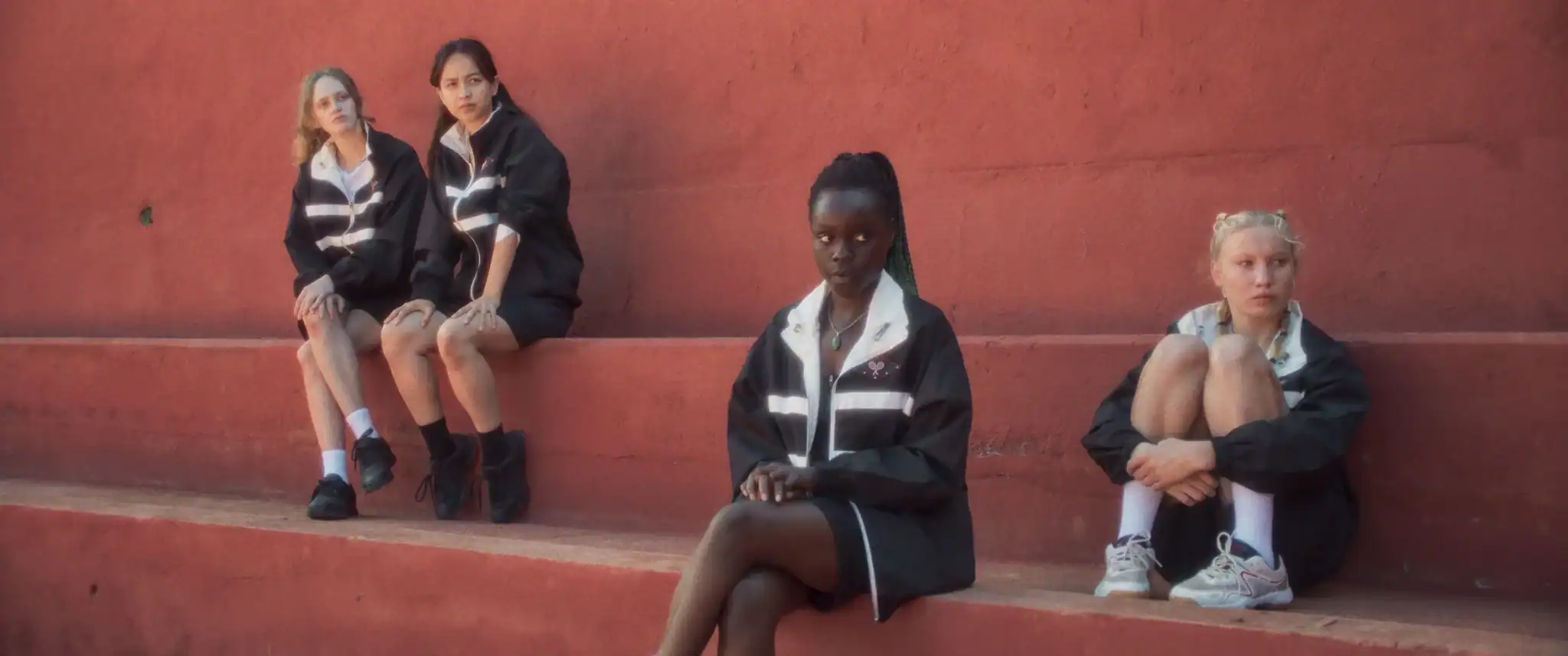 Four young women in matching black jackets sit spaced apart on red concrete steps, looking in different directions.