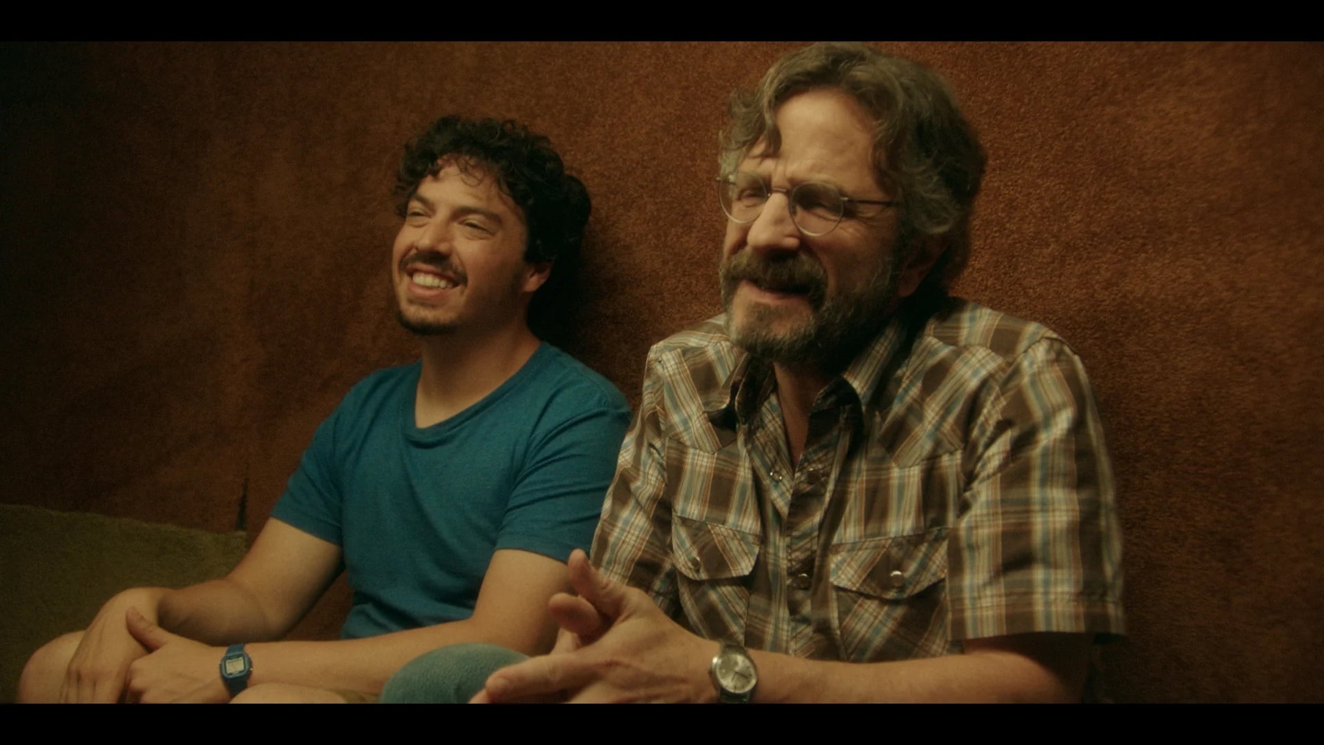 Two men sit next to each other against a brown wall. One wears a blue shirt and smiles, the other has glasses, a plaid shirt, and a watch, appearing to speak or laugh.