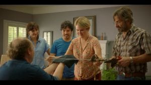 Five adults stand in a living room. One person seated holds a sword, while a standing woman examines it with interest. Others watch and smile.