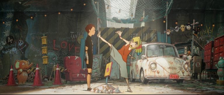 A girl kicks high near a boy in a cluttered garage with graffiti, an old white car, a red chair, and various objects scattered around.